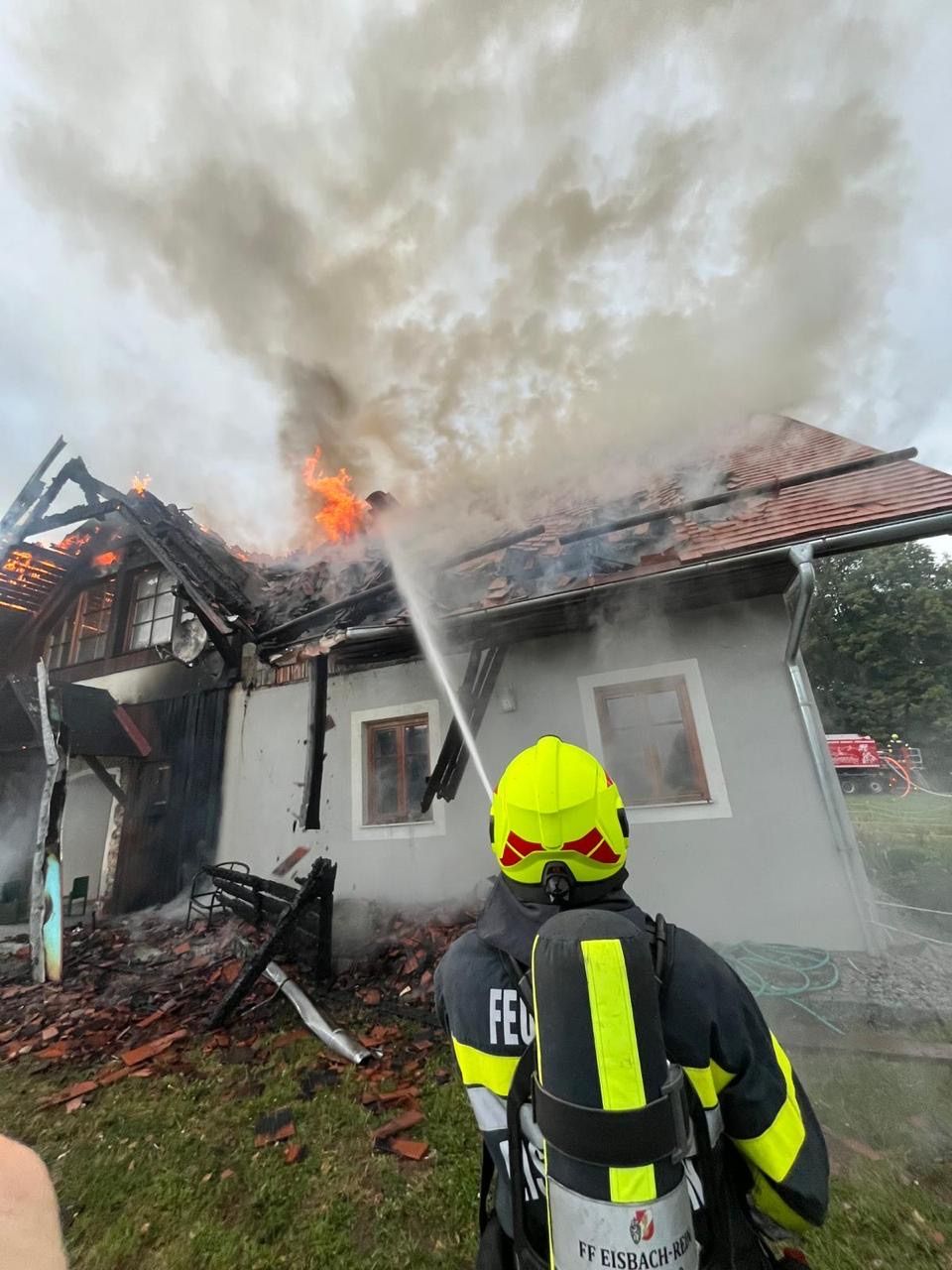 Ein Feuerwehrmann in einem gelben Helm spritzt Wasser auf ein brennendes Haus. Das Haus hat ein beschädigtes Dach und Rauch kommt heraus.