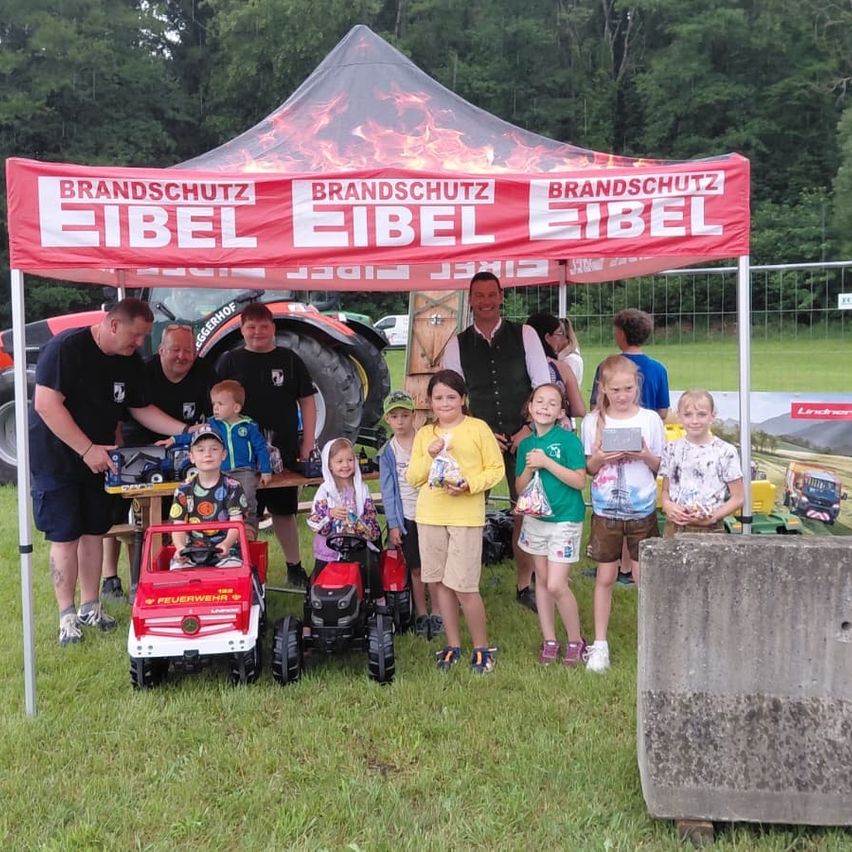 Eine Gruppe von Erwachsenen und Kindern unter einem roten Zelt mit Brandschutz Eibel. Sie haben Spielzeug und posieren für ein Foto.