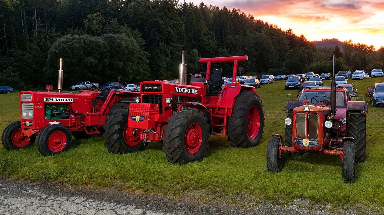 Ein roter Volvo BM Traktor steht auf Gras neben anderen Fahrzeugen. Dahinter zeichnet sich ein Sonnenuntergang am Himmel ab. Bäume säumen das Gebiet, und im Hintergrund sind Autos geparkt.