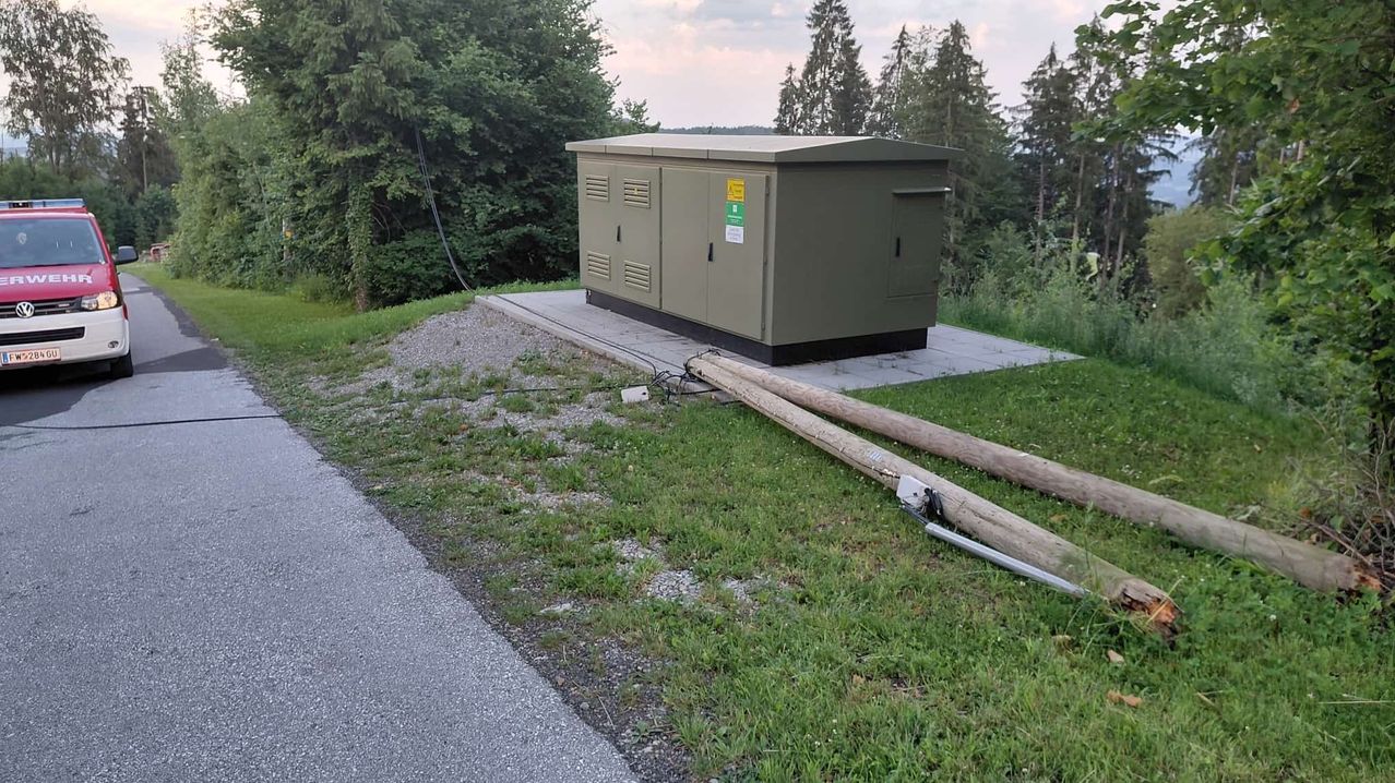 Ein Versorgungskasten mit gelben und grünen Schildern steht auf einem Betonfundament, umgeben von Gras und Bäumen. Zwei Holzpfosten liegen in der Nähe auf dem Gras.