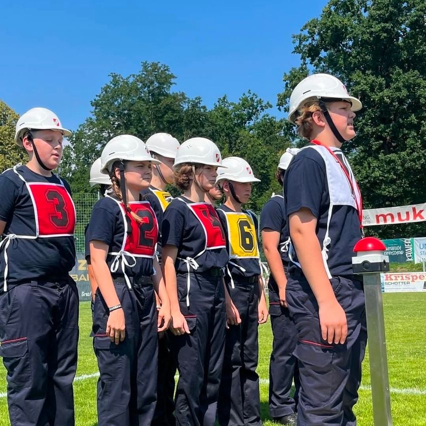 Eine Gruppe junger Menschen in nummerierten Uniformen und Helmen steht auf einem Rasenfeld unter blauem Himmel. Bäume sind im Hintergrund.