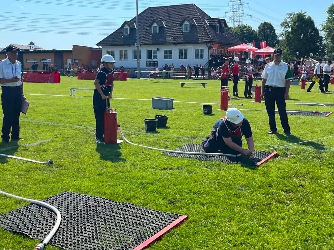 Eine Gruppe von Menschen ist auf einem Feld für einen Wettbewerb versammelt. Eine Person kniet, eine andere steht mit einem Schlauch. Mehrere Eimer und rote Objekte liegen auf dem Boden. Im Hintergrund ein Haus und Menschen unter Schirmen.