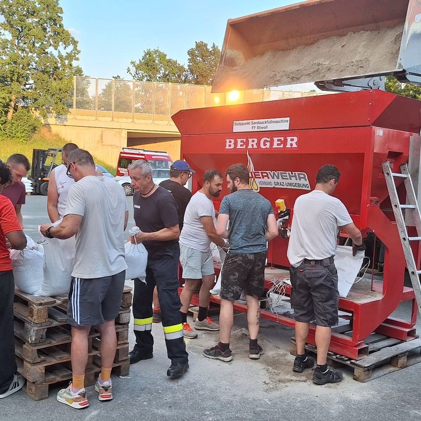 Eine Gruppe von Männern, einige mit Sicherheitswesten, stehen um eine rote Berger-Maschine herum und halten weiße Säcke. Sie scheinen etwas aus der Maschine zu laden oder zu sammeln.
