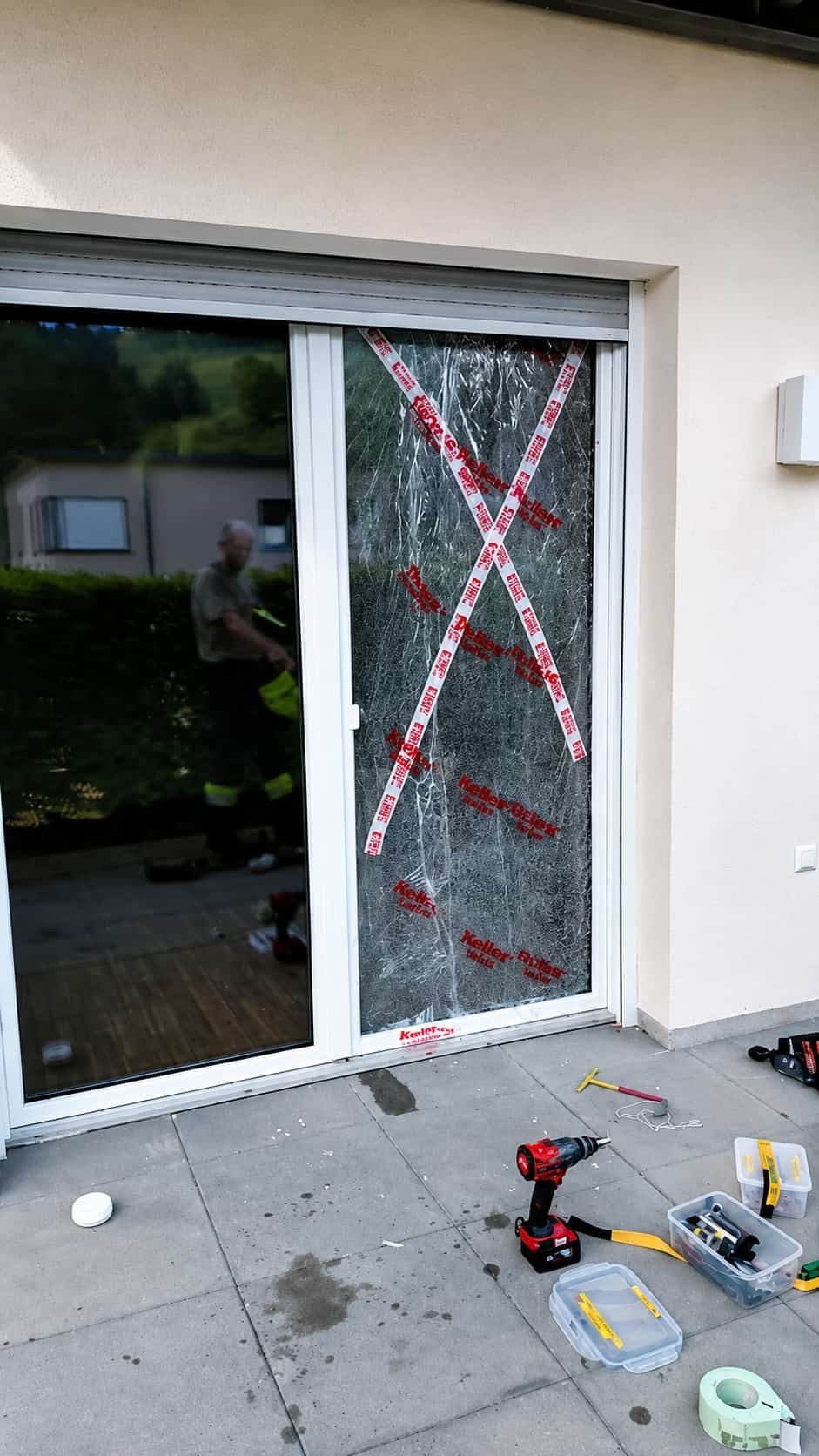 Ein Mann in Sicherheitsausrüstung repariert ein zerbrochenes Fenster mit rotem Klebeband und einem Werkzeug auf dem Boden.