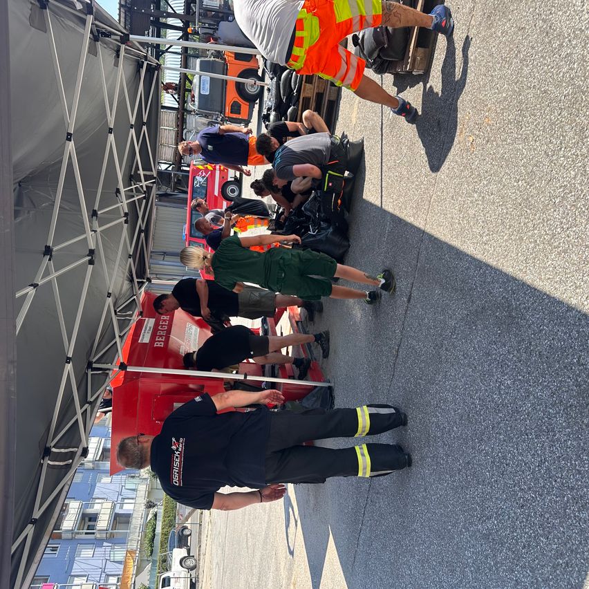 Feuerwehrleute in Uniform arbeiten an einem roten Fahrzeug unter einem Zelt. Einige knien, andere stehen. Im Hintergrund sind Feuerwehrwagen zu sehen.