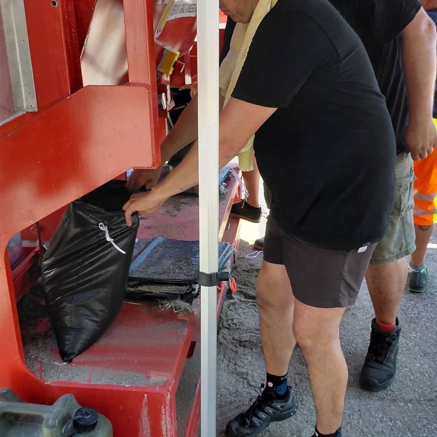 Ein Mann bedient eine rote Maschine und hält einen schwarzen Plastiksack, möglicherweise zum Entsorgen von Abfällen. Ein anderer Mann steht hinter ihm in einer Außenumgebung.