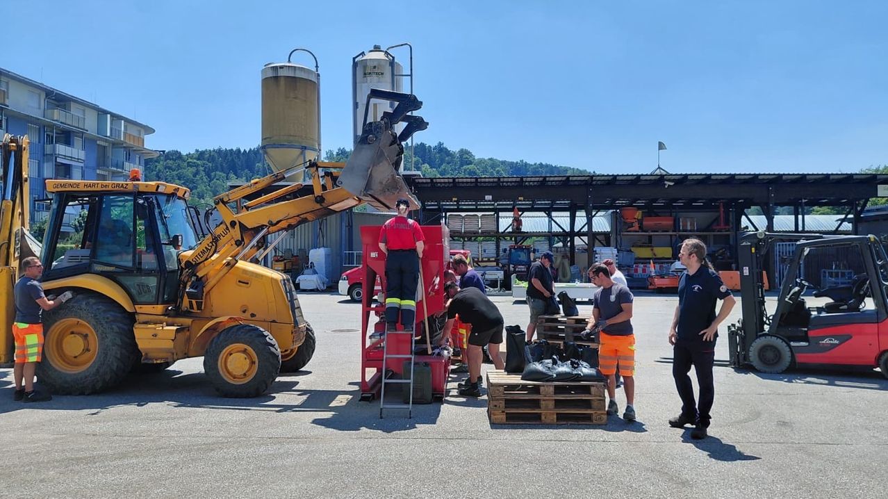 Arbeiter in Schutzausrüstung laden Säcke auf Paletten, mit Maschinen in der Nähe. Ein gelber Bagger und ein rotes Fahrzeug sind vorhanden, zusammen mit mehreren Lagertanks.