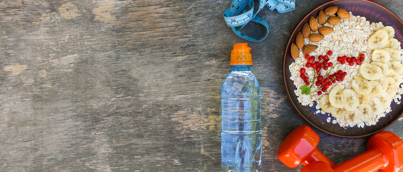 Eine Holzoberfläche mit einem Maßband, einer Wasserflasche, einer Schüssel Frühstück und einer Hantel.