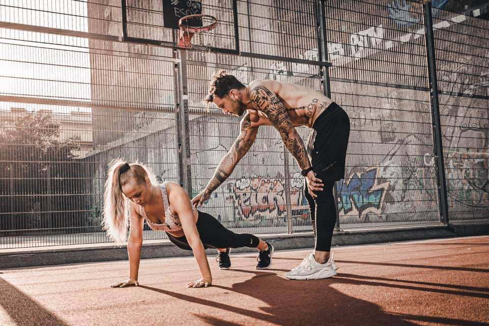 Ein Mann und eine Frau trainieren im Freien. Die Frau macht Liegestütze und der Mann beugt sich hinter ihr. Sie befinden sich auf einem Basketballplatz.