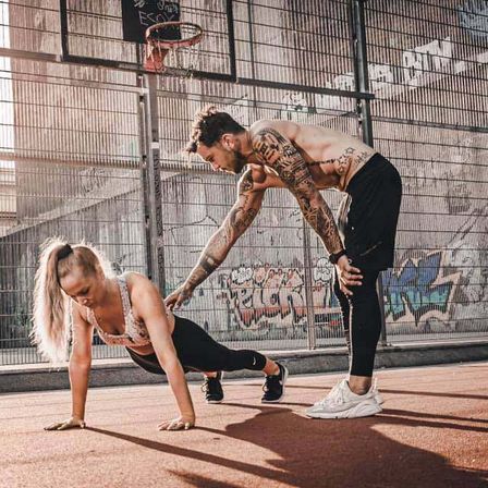 Ein Mann und eine Frau trainieren im Freien. Die Frau macht Liegestütze und der Mann beugt sich hinter ihr. Sie befinden sich auf einem Basketballplatz.