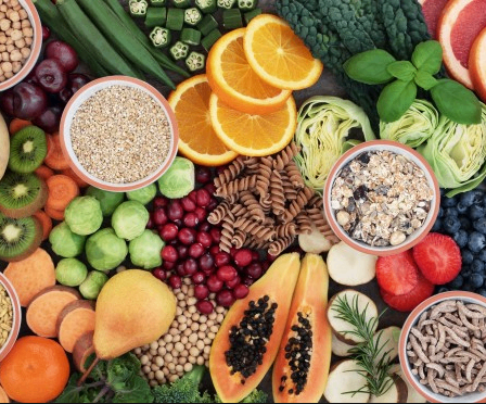 Eine Vielzahl von Früchten, Gemüse, Getreide und Hülsenfrüchten in Schalen und Scheiben, darunter Äpfel, Orangen, Beeren, Brokkoli und Kichererbsen, auf einem Holzuntergrund angeordnet.