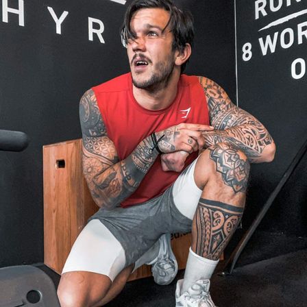 Ein tätowierter Mann sitzt auf einer Holzbank, trägt ein rotes ärmelloses Oberteil und graue Shorts. Er hat ein Mikrofon neben sich und ist in einem Fitnessstudio mit einer schwarzen Wand.