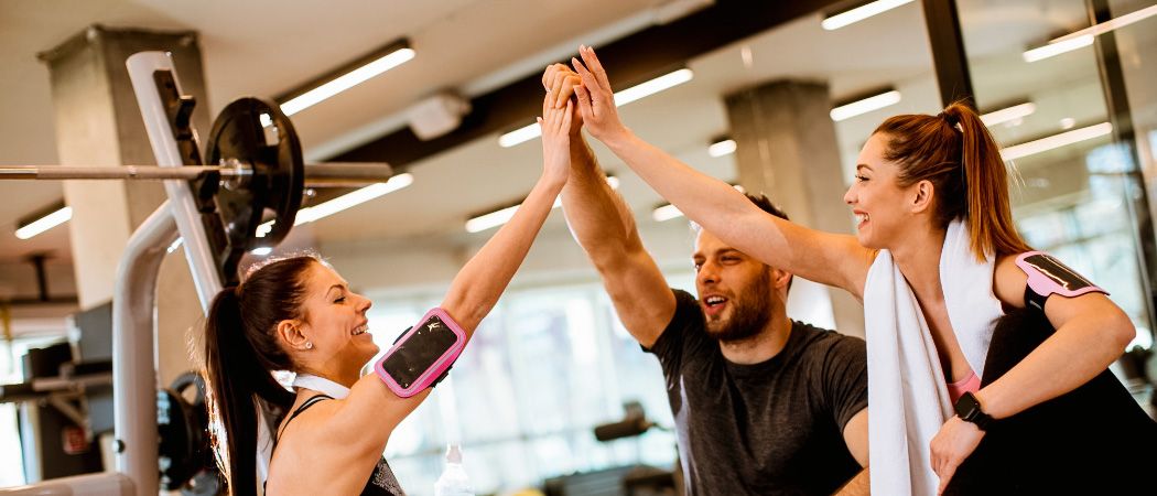Drei Personen in einem Fitnessstudio feiern mit einem High-Five, eine hält einen rosa Fitness-Tracker, eine andere trägt eine Uhr.