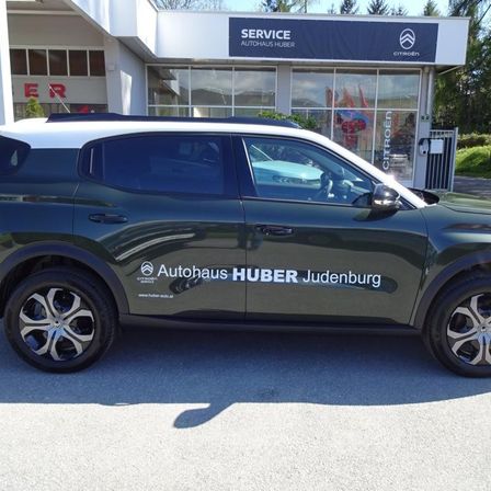 Ein grüner SUV ist vor einem Gebäude geparkt, auf dem ein Schild steht, das 'SERVICE AUTOHAUS HUBER Judenburg' besagt. Der SUV hat ein Logo an der Seite und eine Website-Adresse.