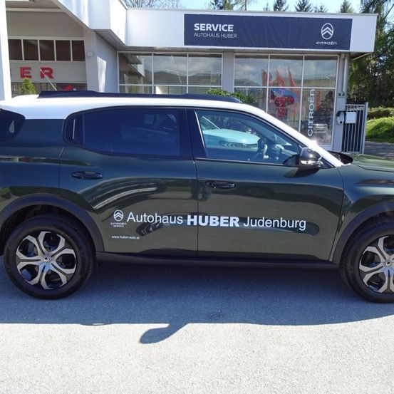 Ein grüner SUV ist vor einem Gebäude geparkt, auf dem ein Schild steht, das 'SERVICE AUTOHAUS HUBER Judenburg' besagt. Der SUV hat ein Logo an der Seite und eine Website-Adresse.