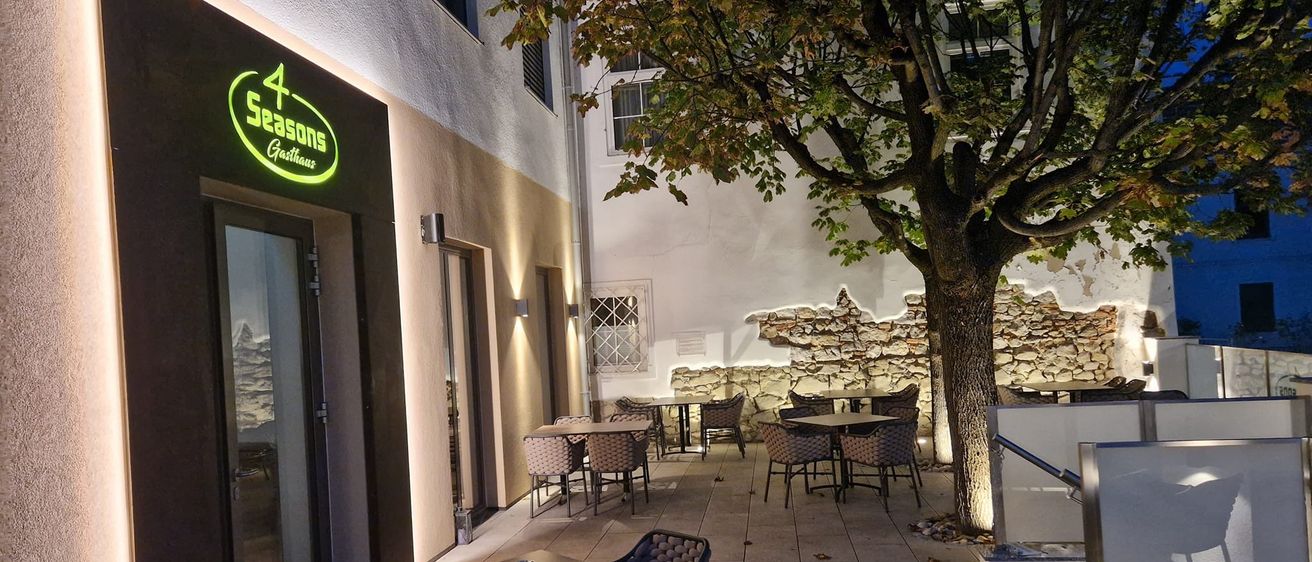 Der Außenbereich eines Restaurants bei Nacht. Tische und Stühle sind um einen Baum herum angeordnet. Das Gebäude hat ein Leuchtschild mit der Aufschrift 4 Seasons Gasthaus.