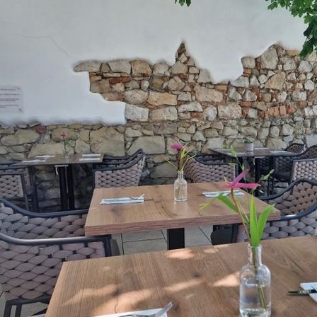 Essbereich im Freien mit Holztischen und Stühlen, einer Steinmauer im Hintergrund und rosa Blumen in Glasvasen.