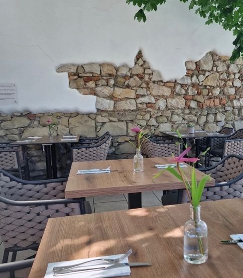 Essbereich im Freien mit Holztischen und Stühlen, einer Steinmauer im Hintergrund und rosa Blumen in Glasvasen.