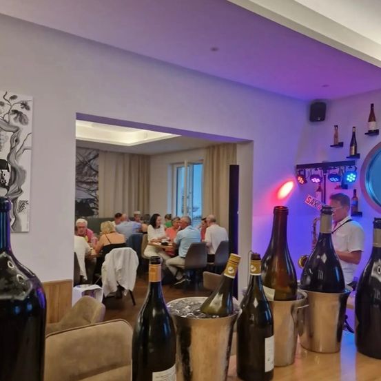 Ein schwach beleuchtetes Restaurant mit einer Bartheke, gefüllt mit Weinflaschen in Eimern. Mehrere Personen sitzen an Tischen, und ein Mann steht mit einer Saxophon.