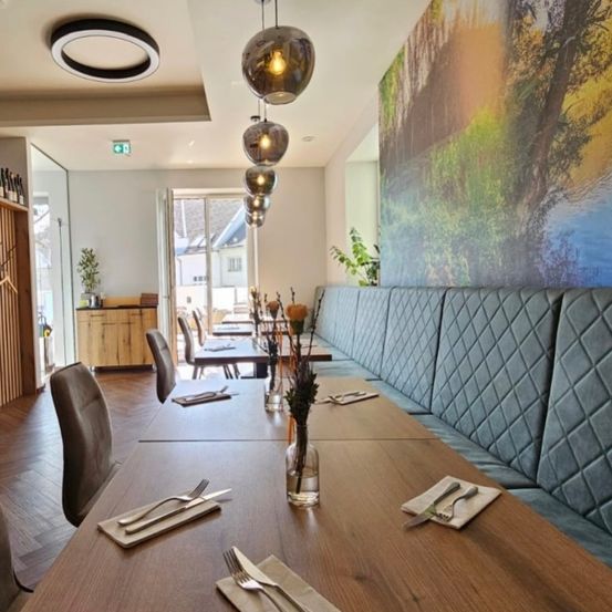 Das Innere eines Restaurants mit einem großen Wandbild, das eine Landschaft darstellt, einem langen Holztisch mit Tischdecken und einer Bank mit blauen Kissen.