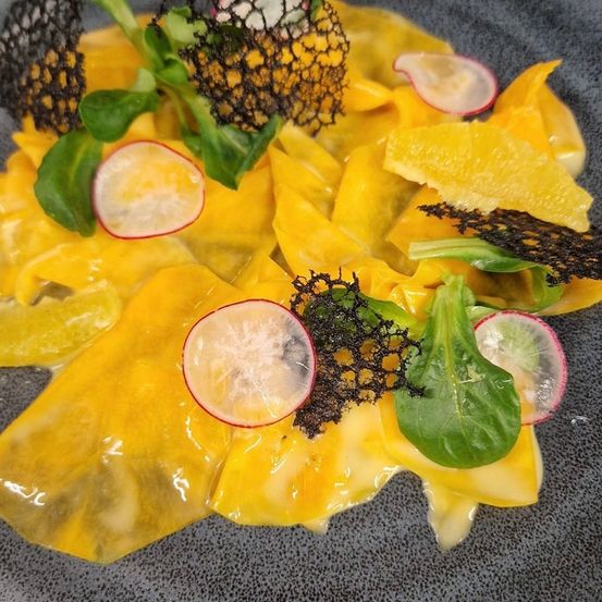 Ein Gericht aus Pasta, möglicherweise Ravioli, mit leuchtend gelber Farbe, garniert mit schwarzen und grünen Gemüsesorten und Radieschenscheiben, auf einem grauen Teller.