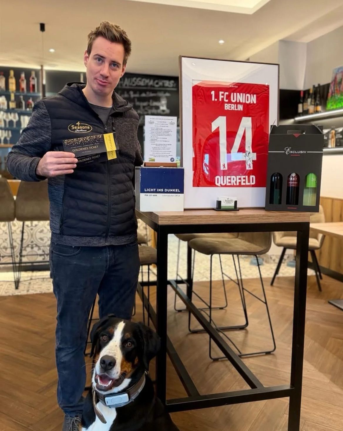 Ein Mann steht neben einem Tisch mit einem signierten Trikot von 1. FC Union Berlin, Nummer 14, zusammen mit einem goldenen Ticket, einer Schachtel Querfeld Zigarren und einem Hund.