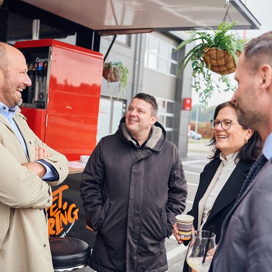 Drei Menschen stehen vor einem Café und lächeln. Ein Mann mit Bart steht links, ein Mann in einem dunklen Mantel in der Mitte und eine Frau in einem schwarzen Mantel rechts. Ein Kaffeeautomat ist im Hintergrund zu sehen.