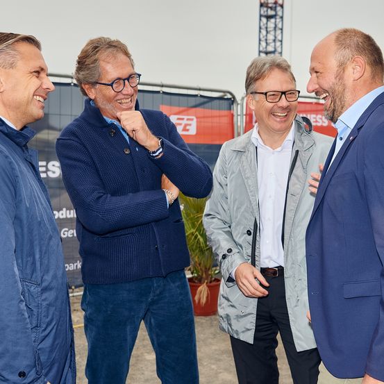 Vier Männer in Businesskleidung stehen lächelnd zusammen draußen. Hinter ihnen ist eine Kulisse mit roten und weißen Bannern.