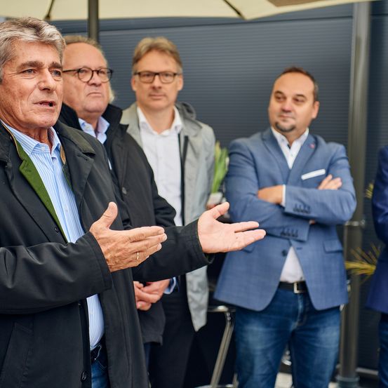 Eine Gruppe von Männern steht in einem Außenbereich. Ein älterer Mann gestikuliert, während er spricht. Hinter ihm stehen zwei Männer mit verschränkten Armen. Sie tragen alle formelle Kleidung.