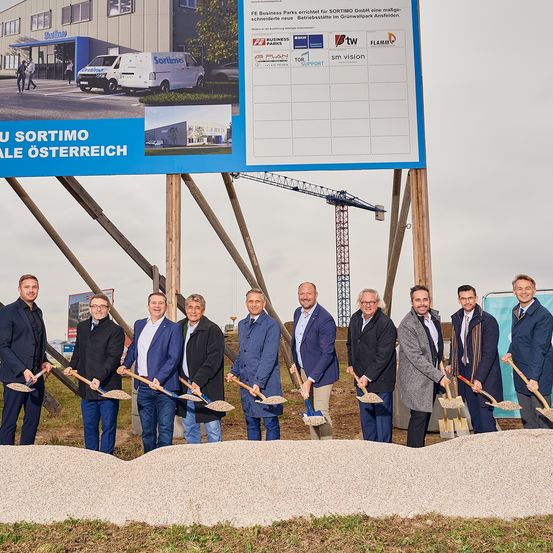 Eine Gruppe von Männern in Anzügen mit Schaufeln posiert für ein Foto vor einem Haufen Erde. Dahinter befindet sich ein großes Banner mit einem Bild eines Gebäudes.