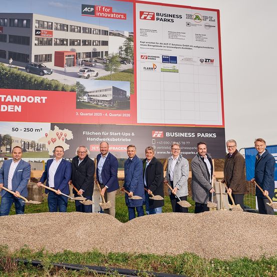 Eine Gruppe von Männern in Anzügen steht vor einem Plakat für Business Parks und hält Schaufeln. Das Plakat zeigt verschiedene Bilder von Gebäuden und eine Karte. Das Gebiet ist mit Erde bedeckt.