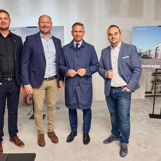 Vier Männer in formeller Kleidung stehen nebeneinander und lächeln für ein Foto. Hinter ihnen befindet sich eine Wand mit einem Banner, auf dem 'Business Parks' steht.