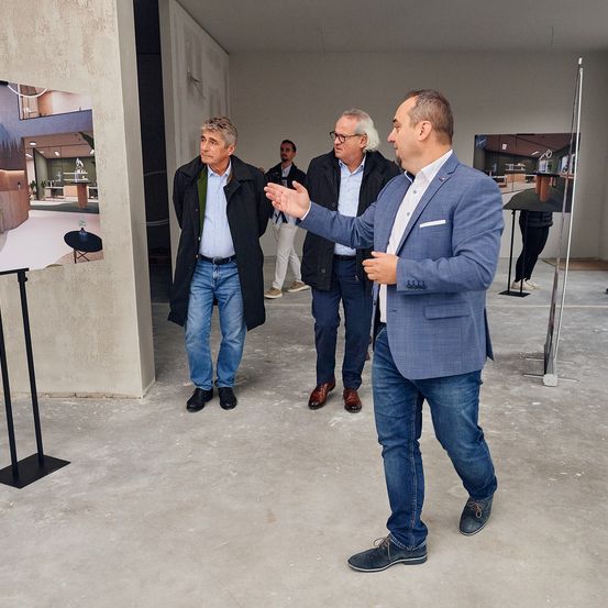 Vier Männer stehen in einem modernen Innenraum. Der Mann im blauen Anzug gestikuliert und spricht. Hinter ihm betrachten zwei andere Männer eine Wand mit Bildern.
