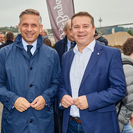 Zwei Männer stehen eng beieinander und lächeln. Sie sind in Anzügen und Krawatten gekleidet. Hinter ihnen stehen Menschen, einige blicken in verschiedene Richtungen. Im Hintergrund befindet sich ein Zaun und ein Gebäude.
