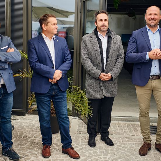 Vier Männer stehen vor einem Gebäudeeingang. Die Männer sind in Anzügen und Lederschuhen gekleidet. Der Mann auf der linken Seite hat die Arme verschränkt, während die anderen lächeln und die Hände vor sich verschränkt haben.