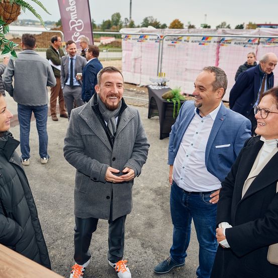 Mehrere Männer und Frauen stehen in der Nähe einer Baustelle. Ein Mann in einem grauen Mantel und orangefarbenen Turnschuhen steht in der Mitte und lächelt. Ein anderer Mann in einem blauen Jackett spricht mit ihm. Im Hintergrund stehen weitere Personen und unterhalten sich in der Nähe eines Tisches mit Pflanzen.
