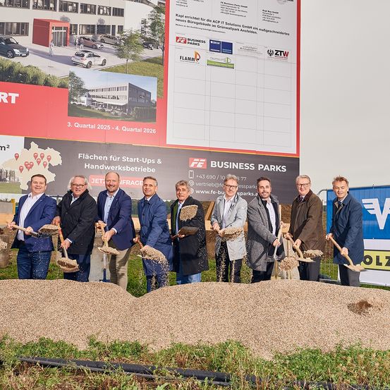 Gruppe von Männern in Anzügen, die Schaufeln vor einer Baustelle eines Gewerbeparks halten. Ein Banner hinter ihnen zeigt 'Business Parks'.