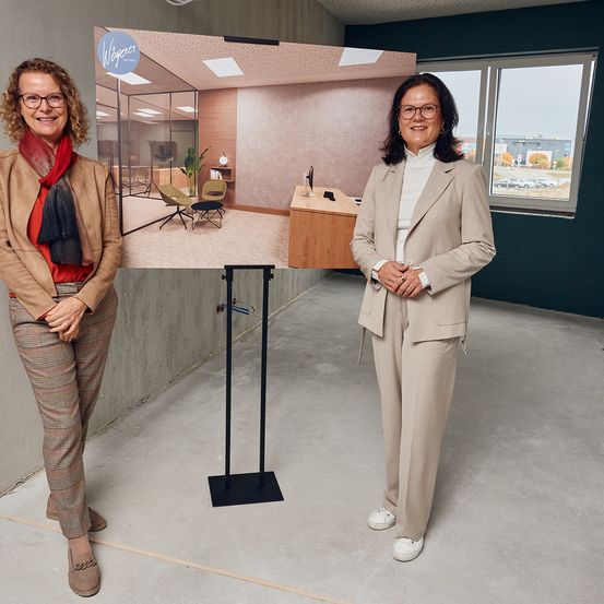 Zwei Frauen in Anzügen stehen in einem Büro. Eine hält ein großes Poster eines Büroraums. Die andere lächelt mit verschränkten Händen.