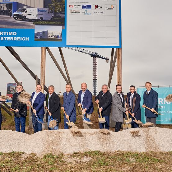 Eine Gruppe von Männern in formeller Kleidung, die Schaufeln halten, steht vor einem Haufen Erde, mit einer Baustelle und einem großen Schild im Hintergrund.