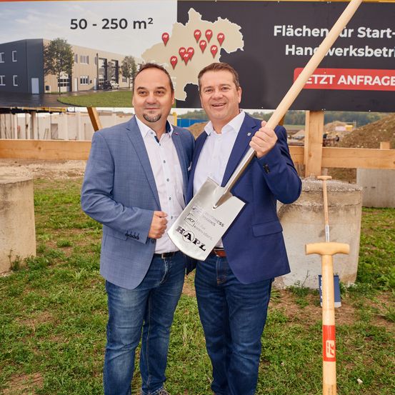 Zwei Männer stehen vor einem Schild mit einer Schaufel, wahrscheinlich an einer Baustelle. Das Schild hat eine Karte und Text über Flächen und Werkstattgrößen.