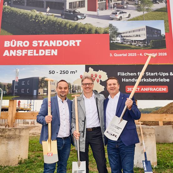 Drei Männer stehen vor einer Baustelle, halten Schaufeln in der Hand und lächeln. Hinter ihnen befindet sich eine große Werbung für 'Büro Standort Ansfelden'. Die Werbung zeigt eine Karte, eine Zeitleiste und eine Einladung zur Anfrage von Informationen.