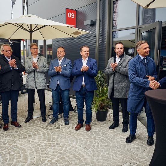 Sieben Männer stehen vor einem Gebäude mit der Nummer 09. Sie klatschen in die Hände.