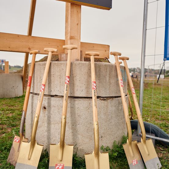 Sechs Schaufeln sind um einen Betonpfeiler herum angeordnet. Jede Schaufel hat einen Holzgriff und eine goldene Klinge. Sie stehen auf einem Grasfeld. Hinter dem Pfeiler befindet sich eine Holzkonstruktion mit einem Schild.