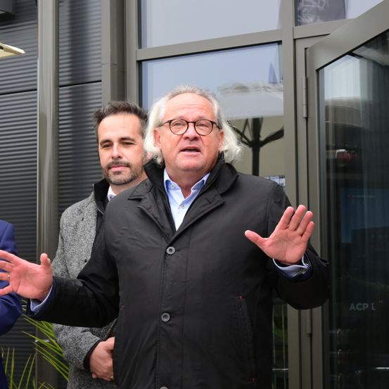 Ein Mann mit weißen Haaren und Brille spricht vor einem Gebäude und gestikuliert. Hinter ihm beobachtet ein Mann in einem grauen Mantel.