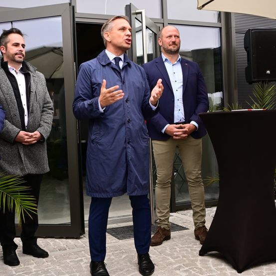 Drei Männer in formeller Kleidung stehen vor einem Gebäude. Der Mann in der Mitte spricht.