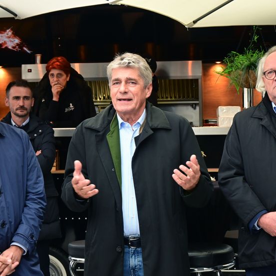 Ein Mann in einem schwarzen Mantel und einem grünen Pullover spricht vor drei Männern, von denen einer eine Brille trägt, in einer Bar mit Theke, Regalen und einer Topfpflanze.