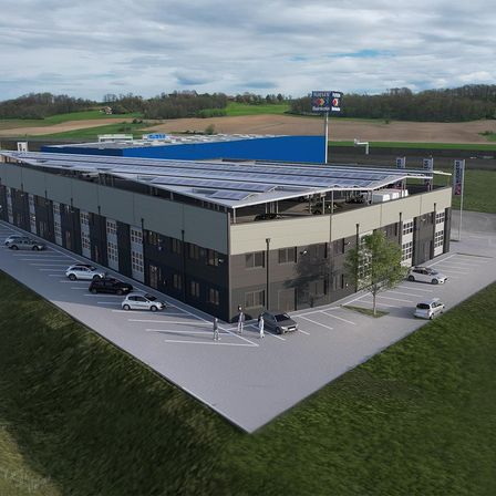 Eine Draufsicht auf ein modernes Gebäude mit Solarpanelen auf dem Dach, umgeben von Autos auf dem Parkplatz. Das Gebäude ist grau mit einem blauen Abschnitt. Bäume und Gras sind im Hintergrund.