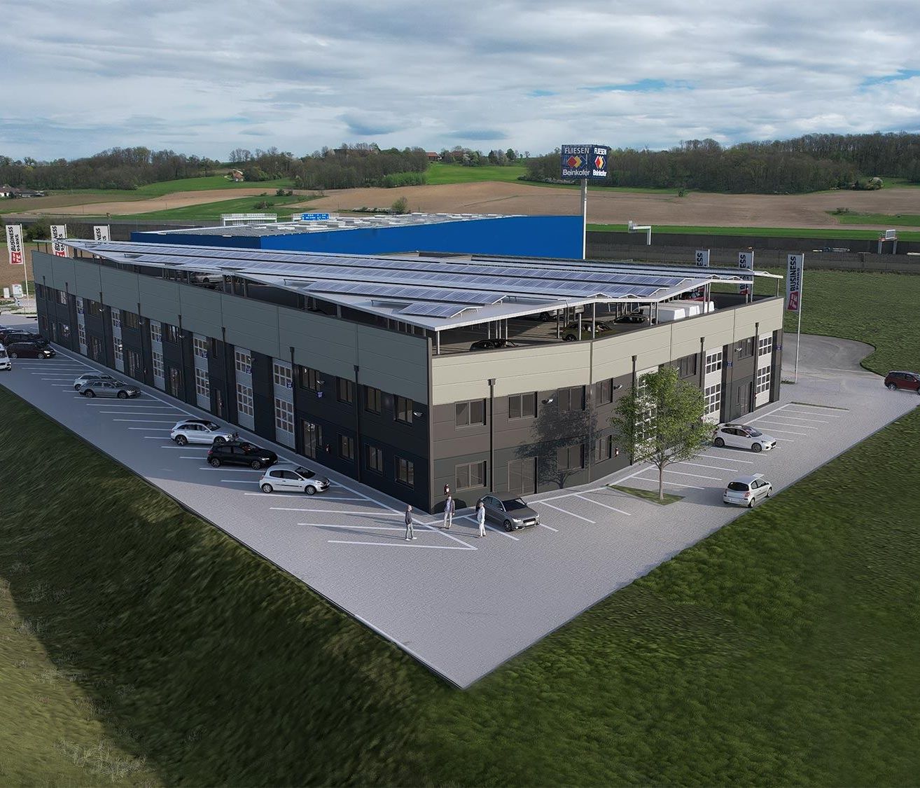 Eine Draufsicht auf ein modernes Gebäude mit Solarpanelen auf dem Dach, umgeben von Autos auf dem Parkplatz. Das Gebäude ist grau mit einem blauen Abschnitt. Bäume und Gras sind im Hintergrund.