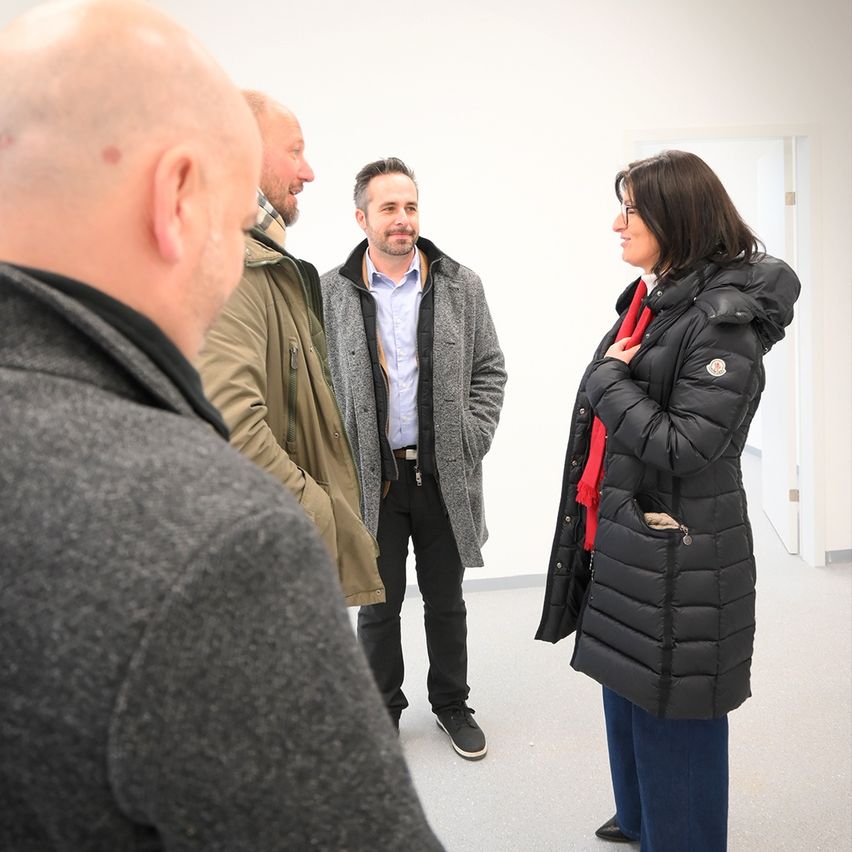 A group of people stand in a room, discussing. A woman in a black puffer jacket and a red scarf is speaking to a man. Behind her, a man in a grey coat looks on.