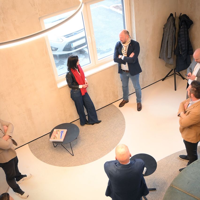 An indoor gathering of people with a woman facing a man. They are standing in a circle with a desk in the middle. A car is parked outside the window.